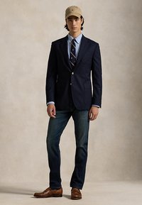 Man draagt een marineblauw colbert, lichtblauw overhemd, een stropdas met patroon, donkere spijkerbroek, bruine schoenen en een beige honkbalpet, staand tegen een neutrale achtergrond.