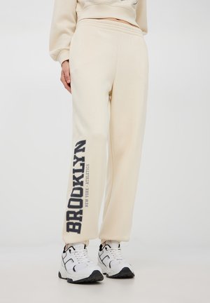 Pantaloni felpa beige con una vestibilità comoda, caratterizzati da una stampa "BROOKLYN" in blu scuro sulla gamba sinistra. Vita elastica e polsini.