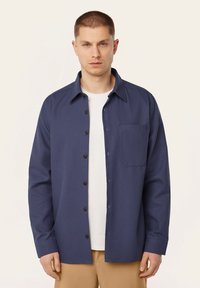 Navy button-up shirt van textiel met textuur, met een borstzak, kraag en lange mouwen; gedragen over een wit T-shirt.