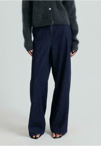 Široke temne kavbojke z visokim pasom, z gladko teksturo in diskretnimi šivi. Ujemajo se s temno puhasto puloverjem.