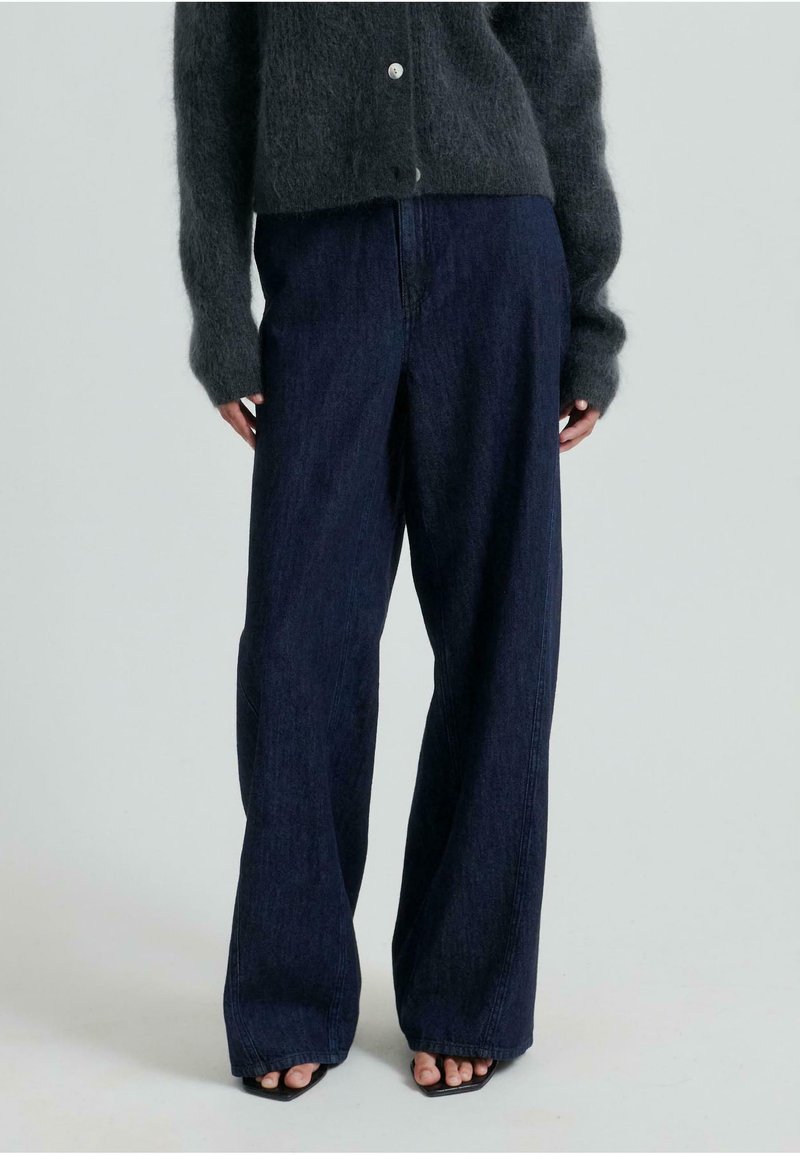 Široke temne kavbojke z visokim pasom, z gladko teksturo in diskretnimi šivi. Ujemajo se s temno puhasto puloverjem.