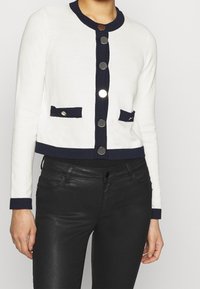 White knit cardigan with navy accents, featuring a round neckline, six metallic buttons, and two front pockets. Cropped design.