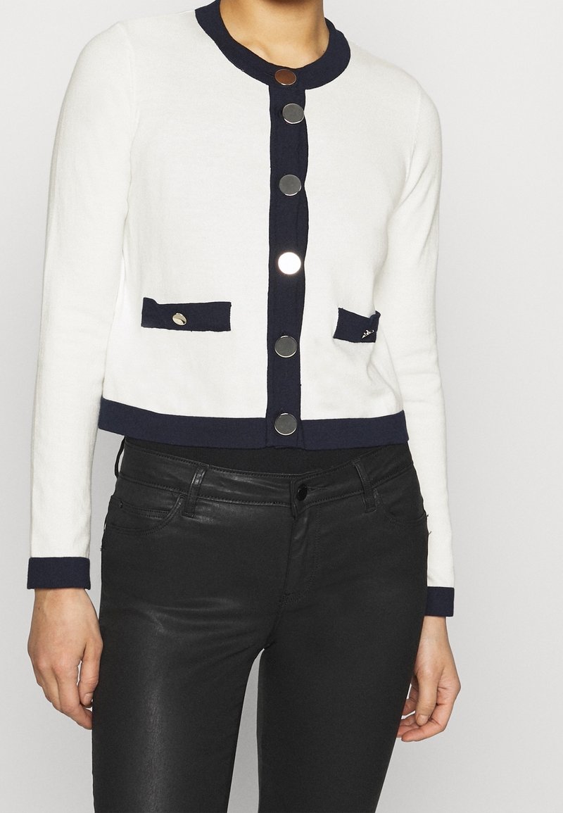 White knit cardigan with navy accents, featuring a round neckline, six metallic buttons, and two front pockets. Cropped design.