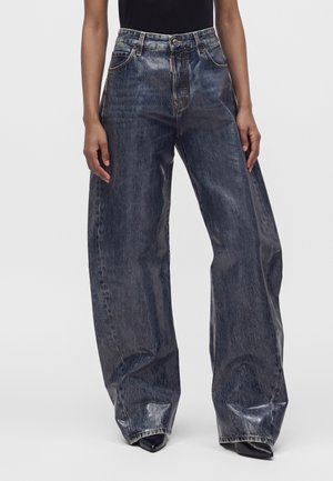 Personne portant un jean taille haute à jambes larges, de couleur foncée avec une finition légèrement brillante, et des chaussures noires pointues visibles sous les ourlets.
