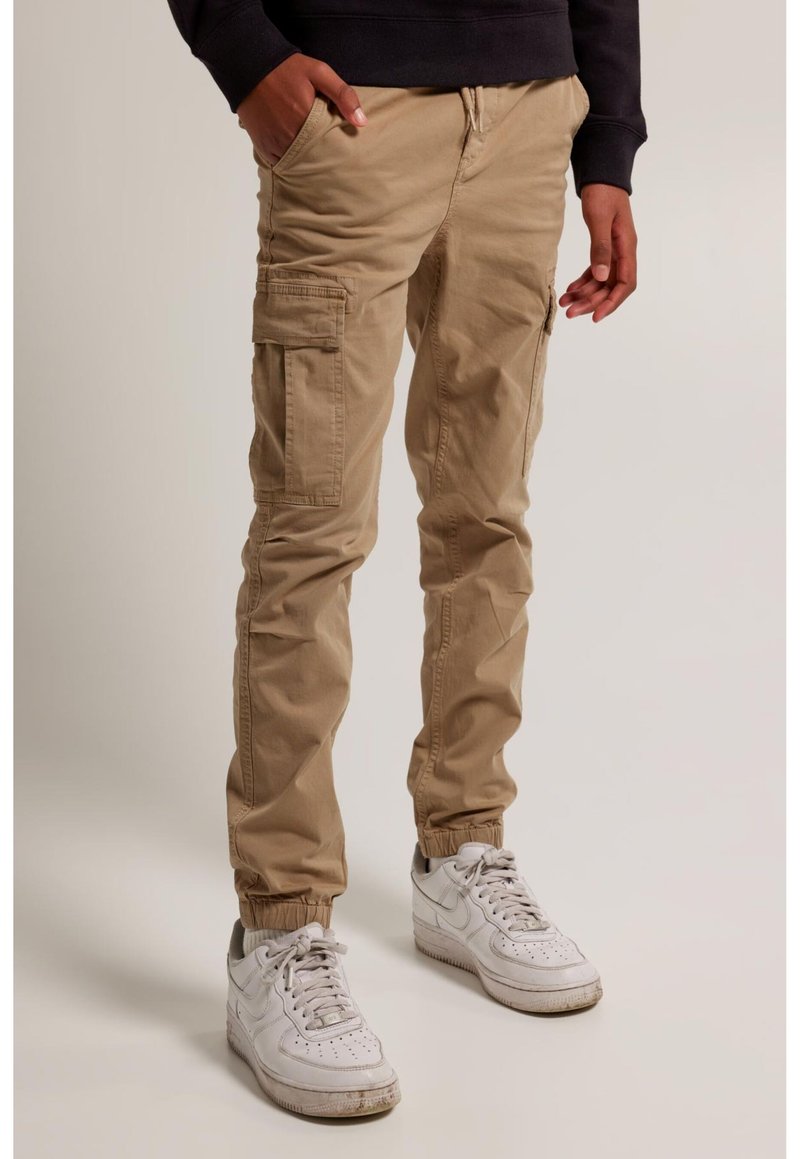 Person trägt beige Cargohosen mit elastischen Bündchen und weiße Sneakers, steht mit einer Hand in der Tasche vor einfarbigem Hintergrund.