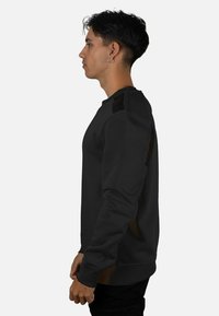 Sudadera negra con cuello redondo, tejido texturizado y detalles en contraste en los hombros. Puños y dobladillo ajustados, vista de perfil lateral.