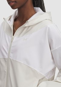 Vit zip-up jacka med huva, med en tvåtonad design. Tillverkad av mjukt tyg har den en subtil Under Armour-logotyp på bröstet.