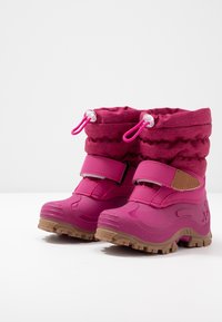 Par de botas de invierno infantiles de color rosa brillante con correas ajustables, parte superior con cordón elástico y suelas texturizadas marrones sobre una superficie blanca.