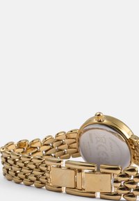 Zlatna kovinska zapestnica s povezanim pasom. Okrogel ohišje z prozornim hrbtom, krono in poliranimi detajli.