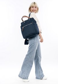 Ung kvinde med kort blondt hår iført hvid top, jeans med vide ben, hvide sneakers, bærer en marineblå rygsæk over skulderen og smiler sidelæns.