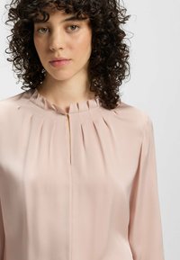 Blusa rosa pallido realizzata in tessuto morbido con scollatura plissettata e apertura a chiave, dotata di maniche lunghe e vestibilità comoda.