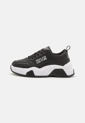 Baskets Versace Jeans Couture noir et blanc avec semelle épaisse, lacets noirs, et texte de marque sur le côté, la languette et la bride de talon.