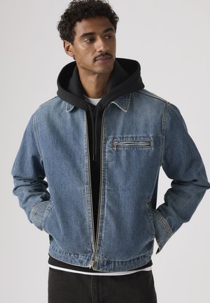 Homme aux cheveux bouclés portant une veste en jean zippée par-dessus un sweat à capuche noir, debout les mains dans les poches, regardant légèrement sur le côté.