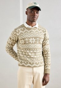 Maglione lavorato a maglia beige con motivi bianchi geometrici, scollo rotondo e polsini a costine. Abbinato a pantaloni color crema e un berretto beige.