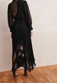 Jupe plissée noire avec superposition transparente et détail de dentelle à l'ourlet, associée à un blouse noire à manches bouffantes et poignets boutonnés.