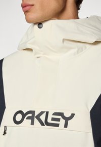 Beige og sort jakke med høj krave; har et fremtrædende sort "OAKLEY" logo, lynlåslomme og mat finish stof.