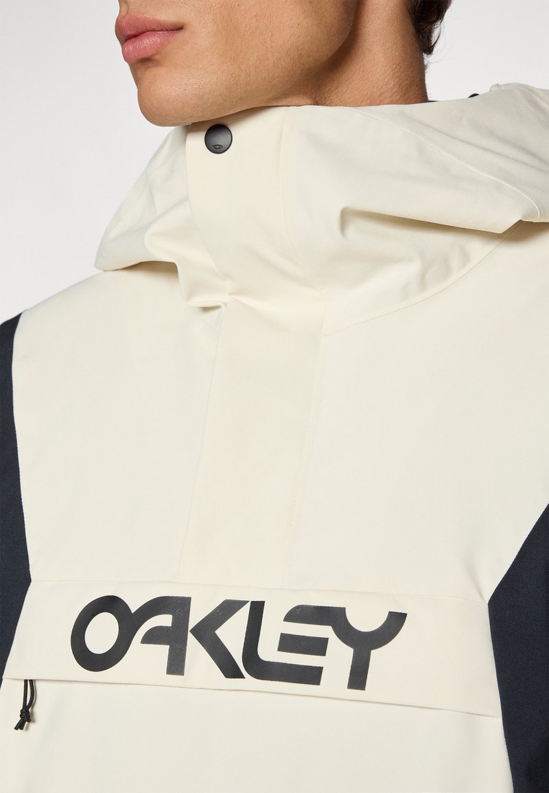 Veste beige et noire avec un col haut ; présente un logo "OAKLEY" noir proéminent, une poche zippée et un tissu au fini mat.
