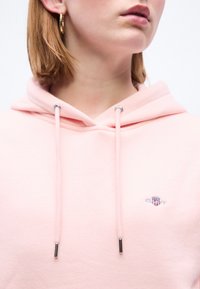 Γυναίκα που φοράει ανοιχτό ροζ φούτερ με κορδόνια και μικρό κεντητό λογότυπο στο στήθος, με ένα χρυσό κρίκο ως σκουλαρίκι ορατό, αποκομμένη κάτω από τα μάτια.