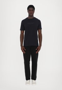 Juoda trumpa rankovė t-shirt su "Armani Exchange" logotipu krūtinėje, derinama su juodomis tiesiomis kelnėmis ir šviesiais sportbačiais.