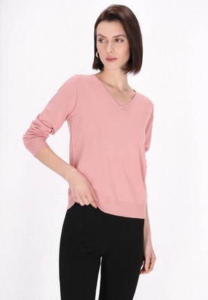 Pulover roz cu decolteu în V, confecționat dintr-un material moale, cu mâneci 3/4 și tiv cu nervuri, asortat cu pantaloni slim negri, pe un fundal alb.