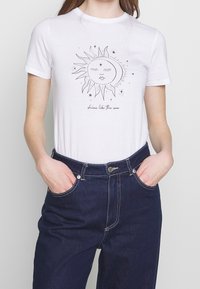 Camiseta de algodón blanca de manga corta con un gráfico de sol negro y el texto "brilla como el sol" en el centro. Combinada con jeans oscuros.