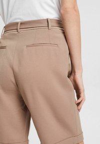 Pantalones cortos marrones a medida con una textura suave, que cuentan con dos bolsillos traseros y puños enrollados, sujetados a la cintura con un cinturón a juego.