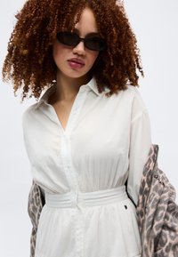 Žena s kovrčavom crvenom kosom koja nosi crne ovalne sunčane naočale, bijelu haljinu stegnutu u struku i jaknu s leopardovim uzorkom prebačenu preko ramena.