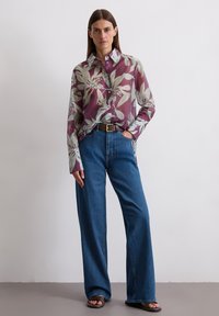 Long-sleeve, button-up shirt with a purple and green floral pattern, paired with loose-fitting blue denim jeans and black sandals.