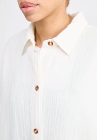 White button-up shirt made of textured fabric, featuring a soft collar and brown buttons along the front, showing subtle vertical striped pattern.