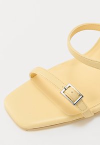 Yellow flat sandal featuring a squared toe, two slender straps with a silver rectangular buckle, and a smooth matte finish.