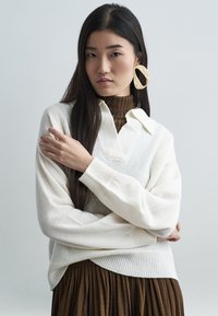 Κρεμ πουλόβερ με γιακά και V-neck, φορεμένο πάνω από έναν καφέ ριγέ ζιβάγκο. Το μοντέλο φοράει κυκλικά σκουλαρίκια, παρουσιάζοντας μια άνετη εφαρμογή.