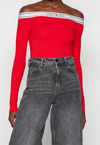 Nainen, jolla on yllään punainen olkaimeton pitkähiha toppi, jossa on valkoinen brändilogo kaula-aukossa, sekä korkeavyötäröiset haalistuneet mustat farkut valkoista taustaa vasten.