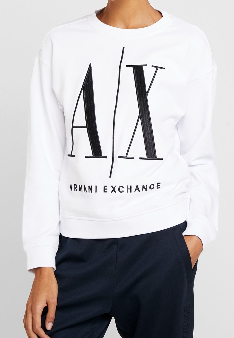Personne portant un sweat-shirt blanc avec de grandes lettres noires "A|X" et le texte "Armani Exchange", porté avec un pantalon noir et une main dans la poche.