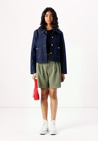 Veste en denim avec un col arrondi, des boutons à l'avant et deux poches poitrine. Portée sur un haut noir, associée à un short vert et des baskets blanches.