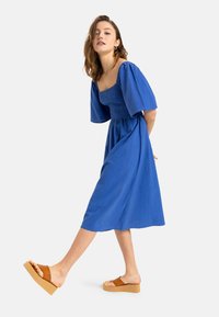 Vestido azul de longitud media con mangas abullonadas y cuerpo fruncido, combinado con sandalias de plataforma marrones. Tejido suave y diseño fluido.