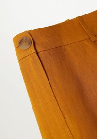 Un pantalon jaune moutarde présente une texture lisse, un seul bouton marron à la taille et un design ajusté aux lignes épurées.
