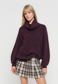 Pull en tricot côtelé violet avec un col cheminée, associé à une jupe plissée à carreaux dans des tons marron, blanc et noir. Manches longues avec poignets ajustés.