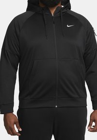 Veste noire zippée à capuche avec poches avant, logo Nike sur la poitrine gauche, et une poche zippée sur la manche gauche, portée par une personne à la peau foncée.