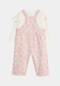 Rosa Latzhose mit weißem Blumenmuster, versehen mit einem Rüschenshirt mit langen Ärmeln. Aus weichem Baumwollstoff, verstellbare Träger mit weißen Akzenten.