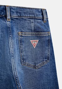 Heledad teksased sinises tooni, millel on oranž õmblus ja kolmnurkne punane-valge "GUESS" logo tagakotil.