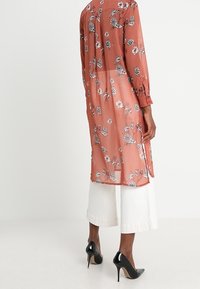 Long, sheer rust-colored tunic with floral prints, buttoned sleeves, and a scooped back hem, paired with white wide-leg trousers and heels.