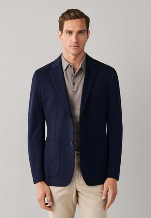 Hombre con blazer azul marino sobre camisa polo gris y pantalones beige, de pie frente a un fondo gris liso.