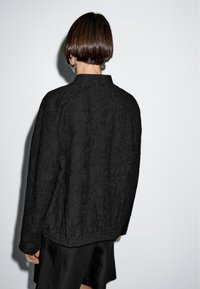 En person med kort brunt hår står vänd mot en vit vägg, klädd i en svart strukturerad jacka och svart kjol, med silverfärgade hoopearrings synliga.