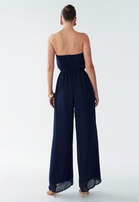 Temno modra kombineza brez naramnic z nabranim zgornjim delom, širokimi hlačnicami, mehko tkanino in rahlo prosojno teksturo na robu.