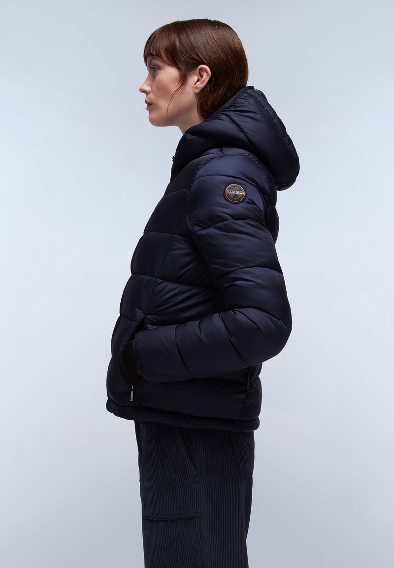 Veste matelassée bleu marine avec capuche, design matelassé, fermeture éclair à l'avant et patch logo sur la manche. Matériau doux et durable avec une finition lisse.