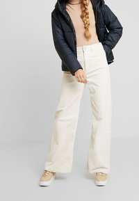 Femme portant une doudoune noire, un col roulé beige, un pantalon large en velours côtelé crème, et des baskets beige, se tenant devant un fond uni.