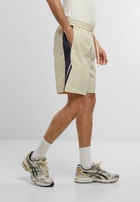 Beige shorts dengan panel samping biru tua, pinggang elastis, dan tali serut. Dipadukan dengan sepatu lari perak dan kaus kaki putih panjang.