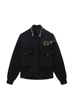 Fekete zipzáras dzseki magas gallérral, két elülső zsebbel és egy arany kígyómintás díszítéssel a bal oldalon. Puha, textúrált anyag.