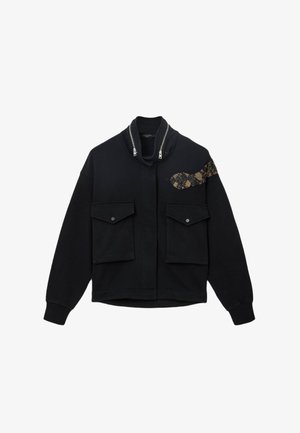 Chaqueta negra con cremallera y cuello alto, dos bolsillos frontales y un detalle de patrón de serpiente dorada en el lado izquierdo. Material suave y texturizado.