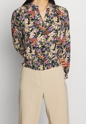 Frau trägt eine langärmlige, schwarze Bluse mit Blumenmuster in Lila, Orange und Creme, kombiniert mit beigen, hoch taillierten Hosen.